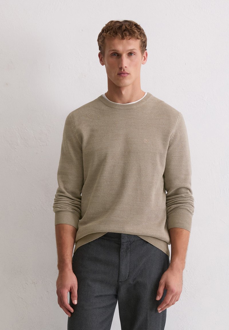 Pull beige texturé avec un col rond et des poignets côtelés, associé à un pantalon gris foncé. Détail logo subtil sur la poitrine.