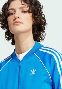 Casaco desportivo azul com fecho éclair, com riscas brancas nas mangas e um pequeno logótipo branco da adidas no peito. Material suave e leve.