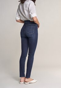 Jeans de tiro alto en azul oscuro con un corte ajustado, con bolsillos traseros y detalles desgastados. Combinados con una camisa de color claro y mules blancos.