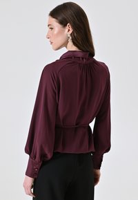 Blusa bordeaux a maniche lunghe con colletto arricciato, vita elastica e polsini con bottoni, realizzata in tessuto liscio.