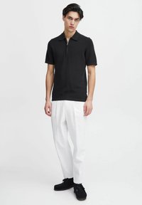 Polo negru cu mânecă scurtă și guler cu fermoar, purtat cu pantaloni din in albi și pantofi slip-on negri, stând pe un fundal simplu.