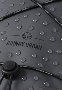 Grått tyg med upphöjda prickar, vattendroppar på ytan, och en svart dragsnodd. Har texten "JOHNNY URBAN" i fet stil.