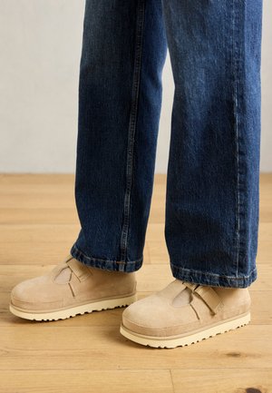 Chaussures en suedine beige avec un design découpé, dotées d'une sangle à velcro et d'une semelle épaisse crème, portées avec un jean en denim bleu ample.