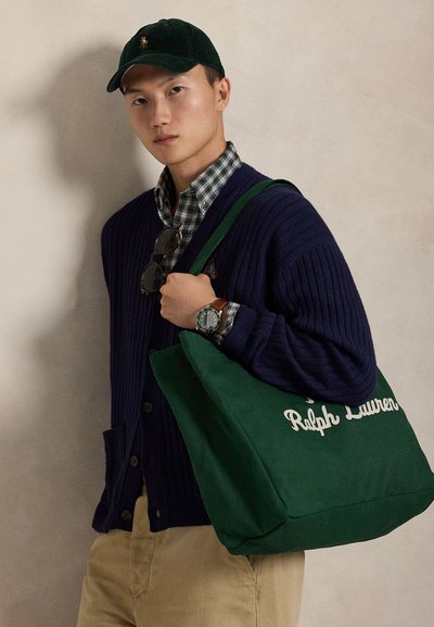 Άνδρας που φοράει σκούφο σκούρου πράσινου, μπλε σκούρο ζακέτα, πουκάμισο καρό, μπεζ παντελόνι, κρατώντας πάνω στον ώμο του μια μεγάλη πράσινη τσάντα Ralph Lauren.