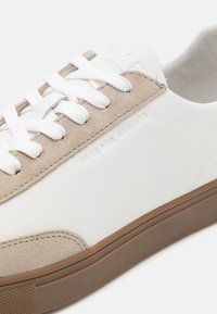 Hvite lær sneakers med beige semsket skinndetaljer, med flate hvite snørebånd, en teksturert såle og subtil merking på siden.
