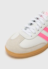Zapatilla blanca con rayas rosas, punta de ante y suela de goma texturizada. Presenta cordones blancos y una lengüeta acolchada.