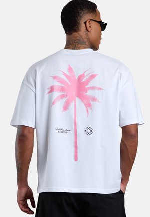 Mann trägt weißes übergroßes T-Shirt mit großem pinkfarbenem Palmenmotiv und kleinem schwarzem Text auf der Rückseite, schwarze Shorts und Sonnenbrille.