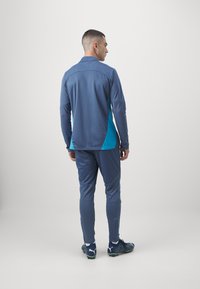 Abbigliamento sportivo composto da una maglietta a manica lunga blu con pannelli laterali turchesi e pantaloni blu coordinati. Tessuto texturizzato e design aderente.