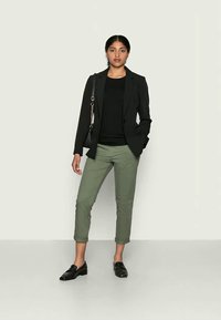 Blazer nero sopra un maglione nero, abbinato a pantaloni verdi oliva tagliati. Mocassini neri completano l'outfit, con una borsa nera in evidenza.
