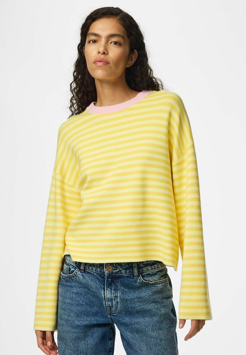 Femme aux cheveux bouclés et foncés portant un pull rayé jaune et blanc avec bordure rose ainsi qu’un jean bleu, debout sur fond blanc.