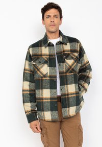 Uomo che indossa una camicia a quadri verde, beige e arancione sopra una maglietta bianca, abbinata a pantaloni cargo marroni, in piedi su uno sfondo bianco.