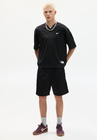 Sort mesh sportsjersey med kortærmer og V-hals, parret med matchende sorte shorts. Tilbehøret omfatter en sølvkæde og lilla sneakers.