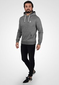 Gray hoodie with a front pocket, drawstring hood, and small logo. Worn with black jeans and black sneakers with gold accents.