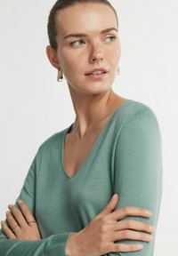 Sweter w serek z miękkiej, turkusowej tkaniny z długimi rękawami. Modelka ma skrzyżowane ręce i nosi przezroczyste kolczyki w kształcie kół oraz delikatne pierścionki.