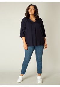 Marineblauwe v-halsblouse met driekwartmouwen, gecombineerd met blauwe skinny jeans en witte sneakers, tegen een neutrale achtergrond.