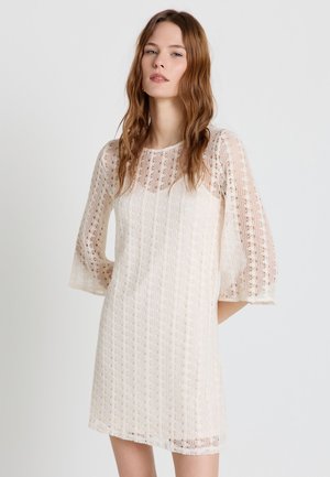 Giovane donna con lunghi capelli castani che indossa un vestito di pizzo bianco lungo fino al ginocchio con maniche a campana ai tre quarti, in piedi davanti a uno sfondo semplice.