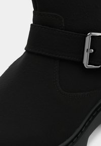 Μαύρο μποτάκι αστραγάλου με υφή στην επιφάνεια, με φαρδιά λουρίδα και ασημένια αγκράφα. Ραμμένες λεπτομέρειες κατά μήκος της σόλας.