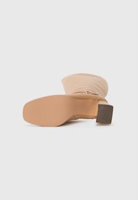 Bottine beige avec une texture lisse, dotée d'un talon bloc et d'une fermeture éclair. La semelle est texturée pour une meilleure adhérence et stabilité.