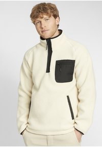 Fleece pullover in cream with a black chest pocket, zip neck, and side inserts. Features a soft texture and a relaxed fit design.