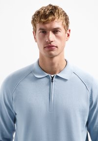 Lichtblauwe pullover met een kwart rits, zachte textuur, puntige kraag en raglanmouwen. Minimale stikseldetails op de schouders.