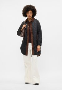 Ženska z afro frizuro nosi prešito črno-rjavo jakno, temno majico, bele široke hlače in drži rjavo torbo preko rame.