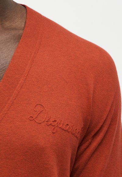 Gros plan sur un pull col V couleur rouille orangé avec un logo "Desigual" brodé de manière subtile sur la poitrine.