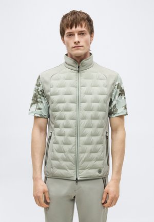 Joven con chaleco acolchado gris claro con cremallera sobre camisa de camuflaje de manga corta y pantalones gris claro, posando frente a un fondo liso.