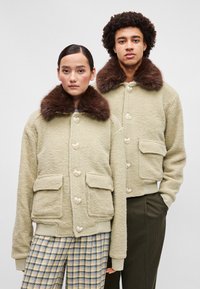 Világos bézs színű bolyhos dzsekik barna műszőrme gallérral. Szív alakú gombok, elülső zsebek és bordázott mandzsetták. Két személy által bemutatva.