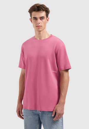 Kurzarm-T-Shirt mit Rundhalsausschnitt in Pink, aus weichem Stoff, mit einem dezenten Logo "PURE PATH" auf der linken Seite, kombiniert mit hellen Jeans.