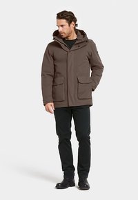 Brun vattentät jacka med huva, två framfickor och en knäppt krage. Matchad med svarta byxor och mörka skor.