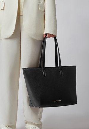 Personne en costume crème tenant un grand sac fourre-tout en cuir noir avec des poignées longues et un petit logo doré au centre inférieur.