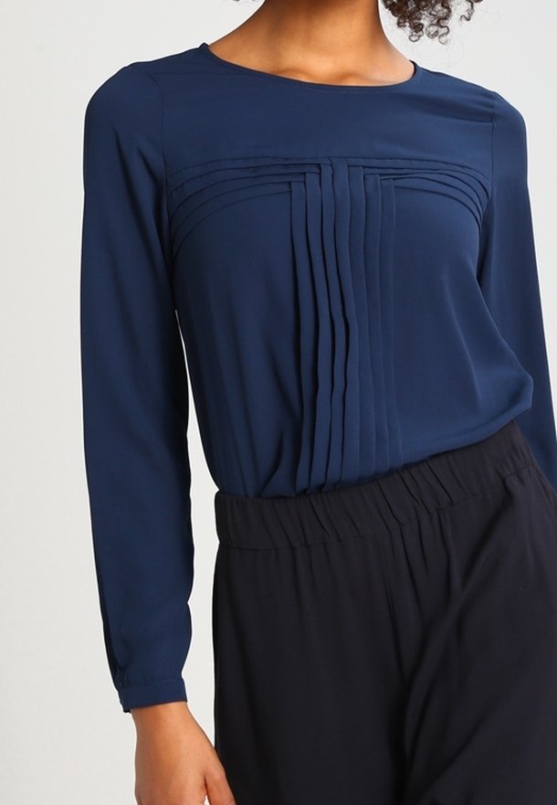 Femme portant un chemisier bleu marine à manches longues avec des plis verticaux sur la poitrine, associé à un pantalon noir taille haute.