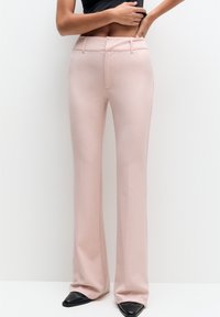 Pantalon rose clair sur mesure en tissu lisse, avec une coupe droite, des passants pour ceinture et une poche avant unique. Chaussures noires à bout pointu.
