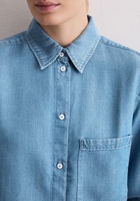 Zbliżenie na osobę noszącą jasnoniebieską koszulę z dżinsu na guziki z widoczną kieszonką na piersiach i kołnierzykiem, częściowo widoczne usta.