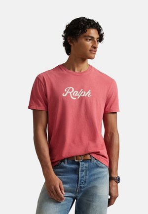 Joven con cabello rizado que lleva una camiseta roja desgastada con la palabra "Ralph", vaqueros azules con cinturón marrón y un reloj de pulsera, mirando hacia su derecha.