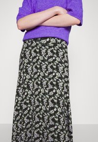 Personne portant un pull court à manches courtes violet et une jupe noire avec un motif floral violet clair, les bras croisés au niveau de la taille.