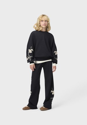 Junge Person mit blondem Haar trägt schwarzen Sweatshirt mit Blumenstickerei und passende weite Hosen mit braunen Schuhen, stehend vor weißem Hintergrund.
