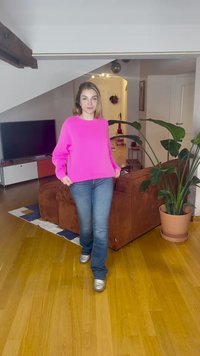 Suéter rosa vibrante com mangas compridas, combinado com jeans de ganga azul e sapatos prateados, numa sala de estar com plantas e móveis.