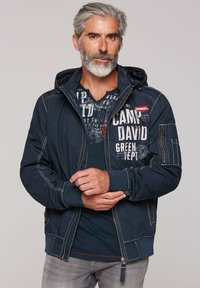 Navy blue hooded jacket with white stitching, featuring a graphic print and zipper pockets. Worn over a dark blue shirt and light gray jeans.