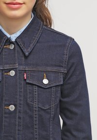 Granatowa kurtka dżinsowa o klasycznym kroju, z kieszeniami na piersi, srebrnymi guzikami, pomarańczowymi przeszyciami oraz miękkim kołnierzem.