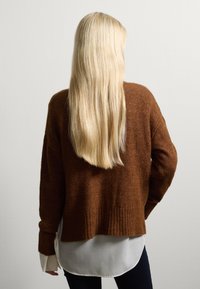 Pull en maille marron à coupe décontractée, avec un ourlet côtelé et des manches longues, superposé sur une chemise blanche, porté avec un jean foncé.