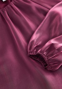 Burgundská saténová látka s hladkou strukturou. Má nařasené elastické manžety a jemné záhyby, které zdůrazňují její tekutý tvar a lesk.
