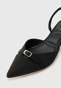 Musta kangas slingback-kengät, joissa on terävä kärki, koristeellinen hihna ja pienet kultaiset solkiaksentit. Teksturoitu kangas ja sileä pohja.