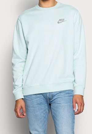 Person trägt hellblauen Nike-Sweatshirt mit Leopardenmuster-Logo auf der Brust und blaue Jeans, steht vor einfachem Hintergrund.