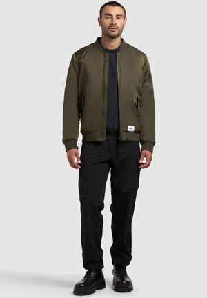 Oliivinvihreä bomber-takki kerroksella ja hihansuilla, vetskari, ja tasku vasemmassa hihassa; yhdistettynä mustiin cargo-housuihin ja mustiin saappaisiin.