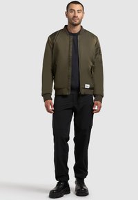 Olivgrüne Bomberjacke mit gestreiftem Kragen und Bündchen, Reißverschluss und einer Tasche am linken Arm; kombiniert mit schwarzen Cargo-Hosen und schwarzen Stiefeln.