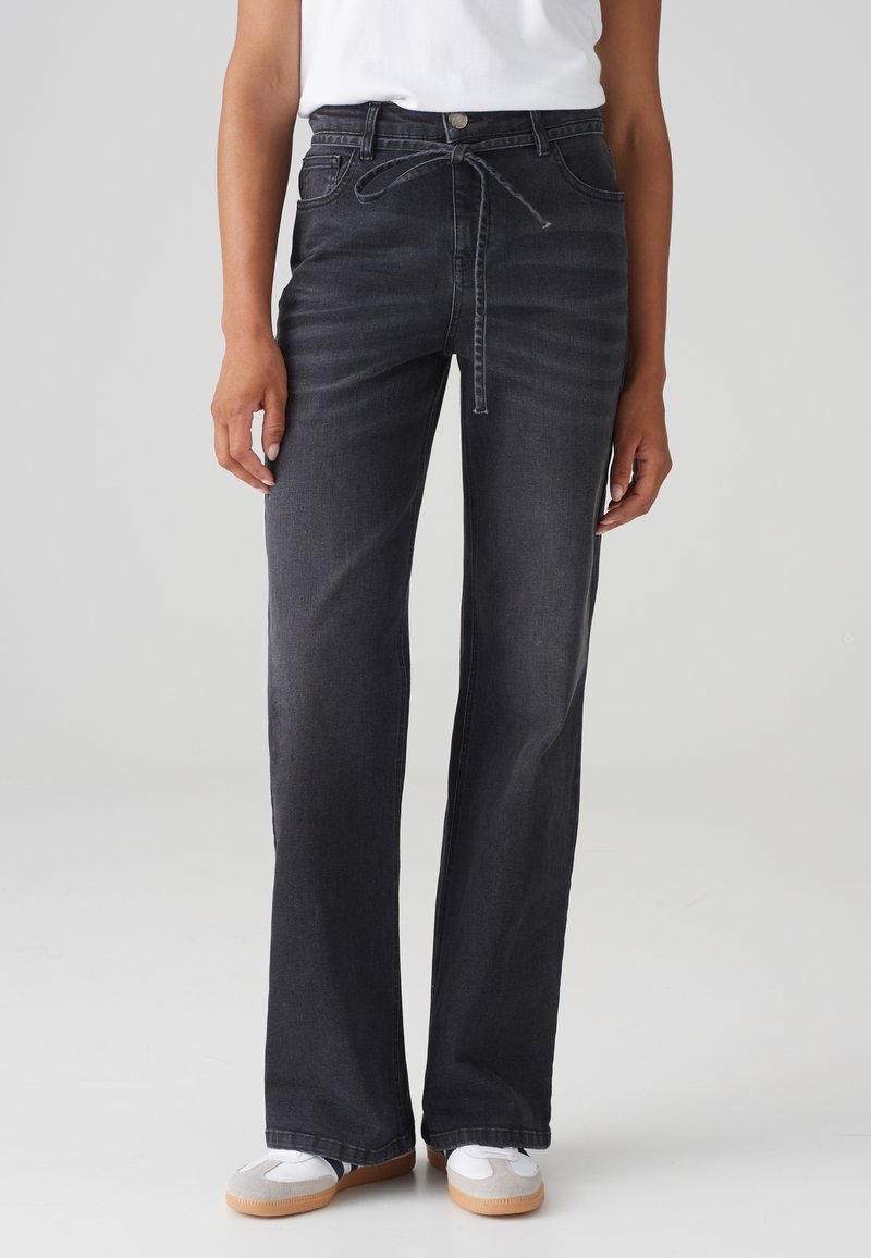 Personne portant un jean noir taille haute à jambes larges avec une ceinture nouée, associé à des baskets blanches et un haut blanc, debout devant un fond uni.