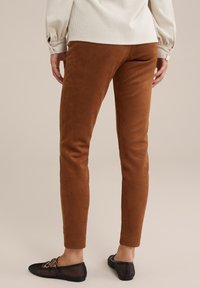 Osoba oblečená v hnědých semišových úzkých kalhotách a černých síťovaných nazouvacích botách stojící před jednobarevným béžovým pozadím.