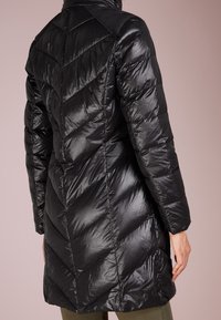 Svart lång quiltad puffjacka med chevron-sömnad som bärs av en stående person mot en enkel ljusrosa bakgrund.