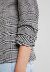 Svart och vit rutigt blazer tyg med röda accenter, strukturerad passform, rullad ärmslut, texturerad med ett hundtandsmönster.